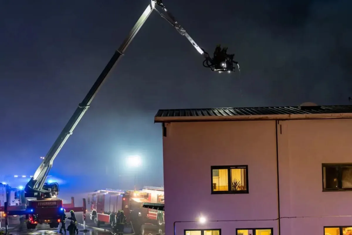 Ein Feuerwehrleiterwagen bekämpft in der Nacht einen Brand auf dem Dach eines Hauses. Einsatzkräfte und Fahrzeuge sind am Boden sichtbar.
