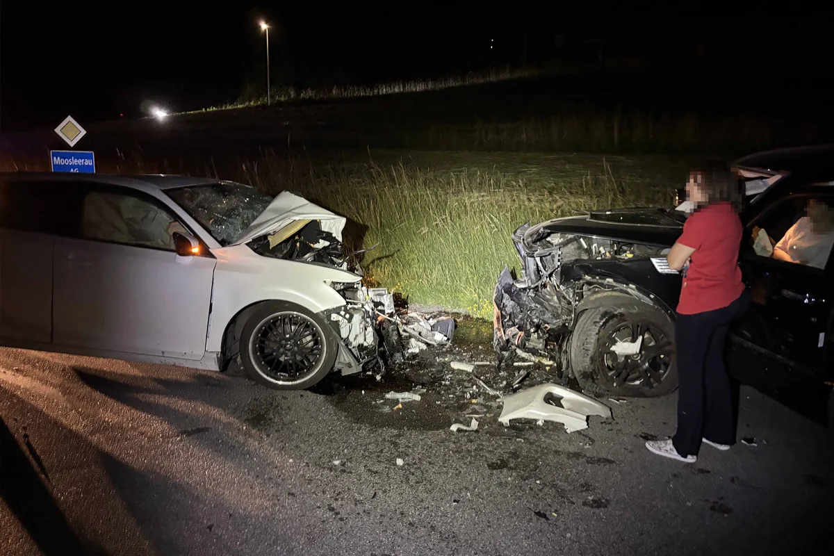 Ein weißes Auto und ein schwarzes Auto stehen stark beschädigt nach einem Frontalzusammenstoß auf der Straße. Eine Person in rotem Hemd betrachtet die Szene.