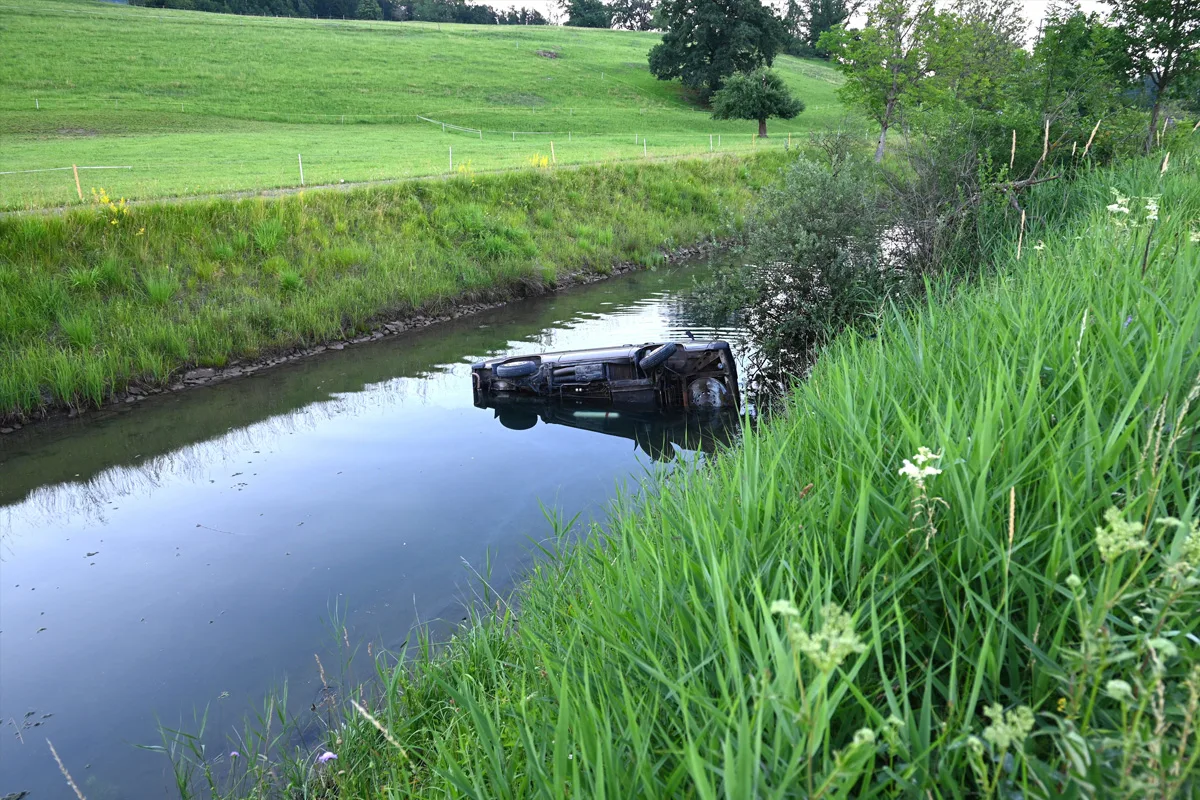 Ein dunkles Auto ist auf der Seite liegend in einem schmalen Wassergraben zu sehen, umgeben von grüner Wiese und Bäumen.