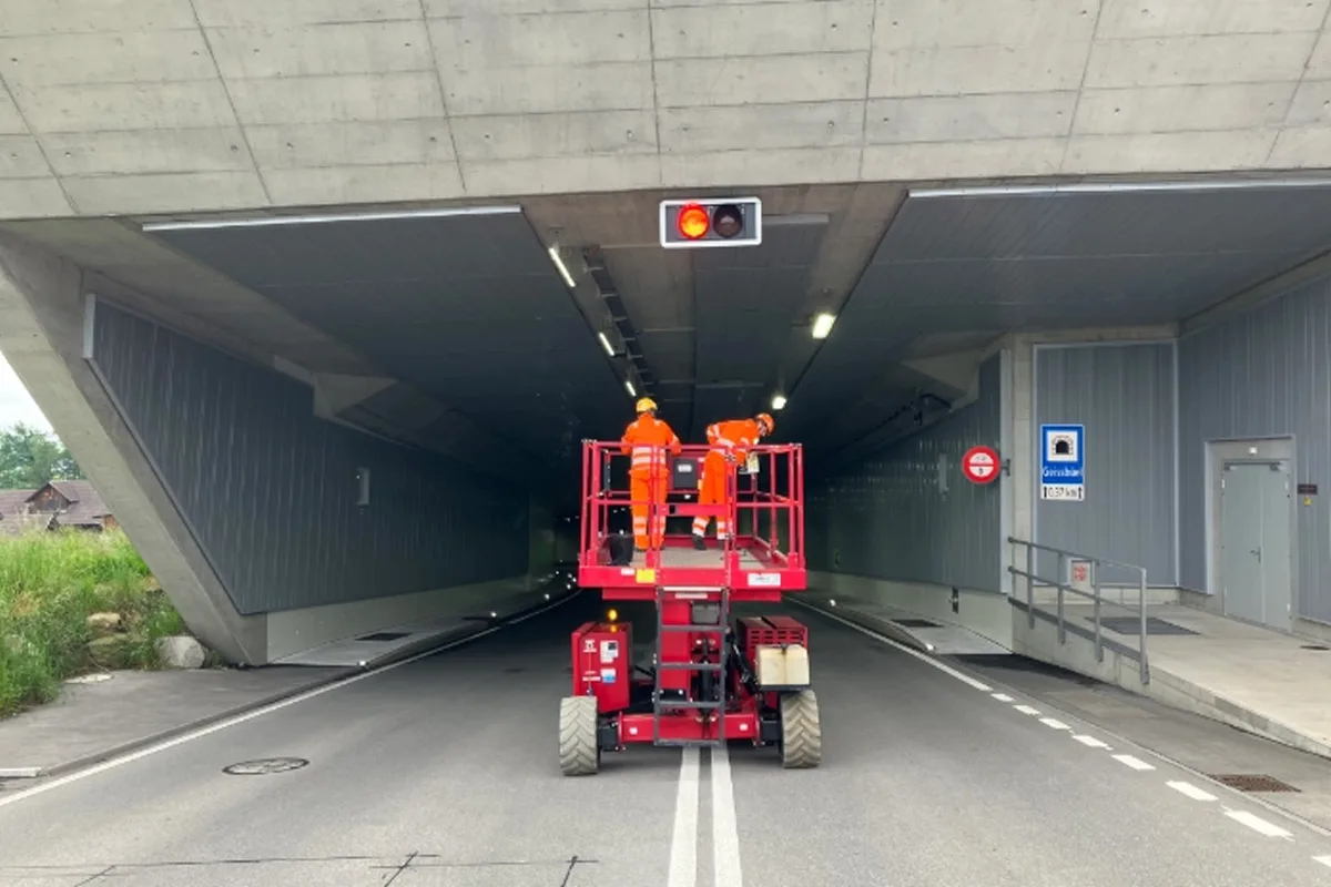 Zwei Arbeiter stehen in Sicherheitskleidung auf einer Hebebühne in einem Tunnel und führen Wartungsarbeiten durch. Der Tunnel ist beleuchtet und eine Umleitung ist eingerichtet.