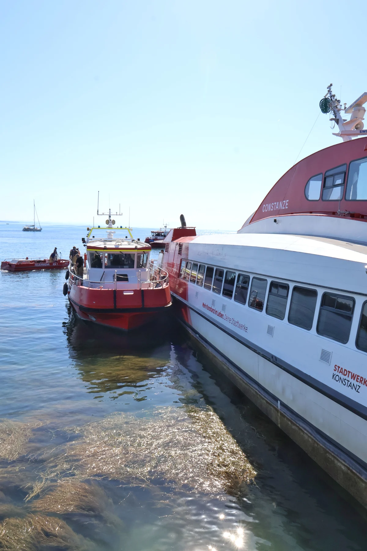Ein Feuerwehrboot und ein größeres Passagierschiff liegen im Wasser nebeneinander. Der Himmel ist klar und sonnig.