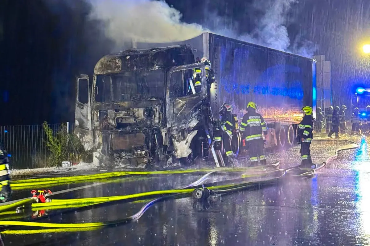 Mehrere Feuerwehrleute in Schutzkleidung löschen einen stark beschädigten und brennenden LKW im Regen. Schläuche und Wasser sind auf der nassen Straße sichtbar.