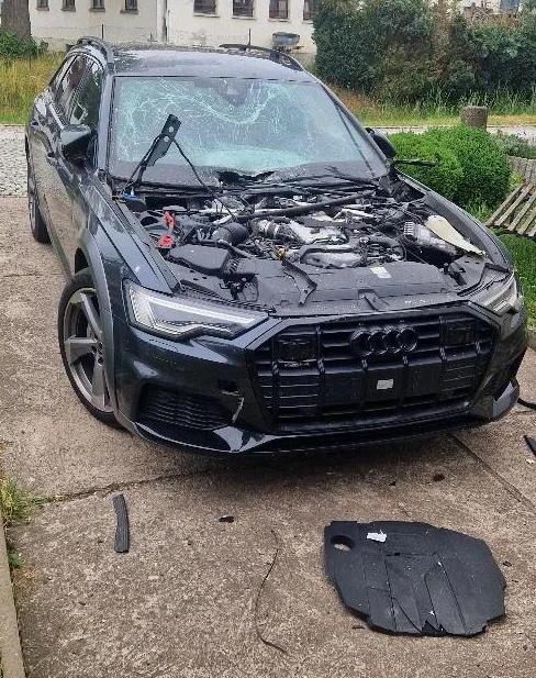 Ein schwarzes Auto steht auf einem Betonhof mit geöffnetem Motorraum und kaputter Frontscheibe. Teile liegen verstreut.