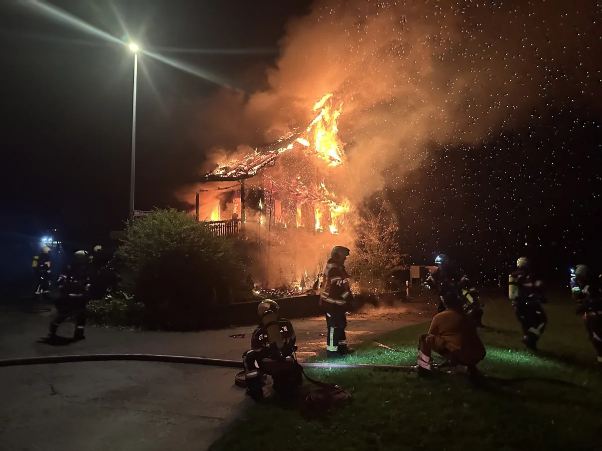Das Bild zeigt ein intensives Feuer, das ein Gebäude erfasst hat, umgeben von Feuerwehrleuten bei Nacht. Die Flammen lodern aus dem Dach, während die Einsatzkräfte davor stehen.