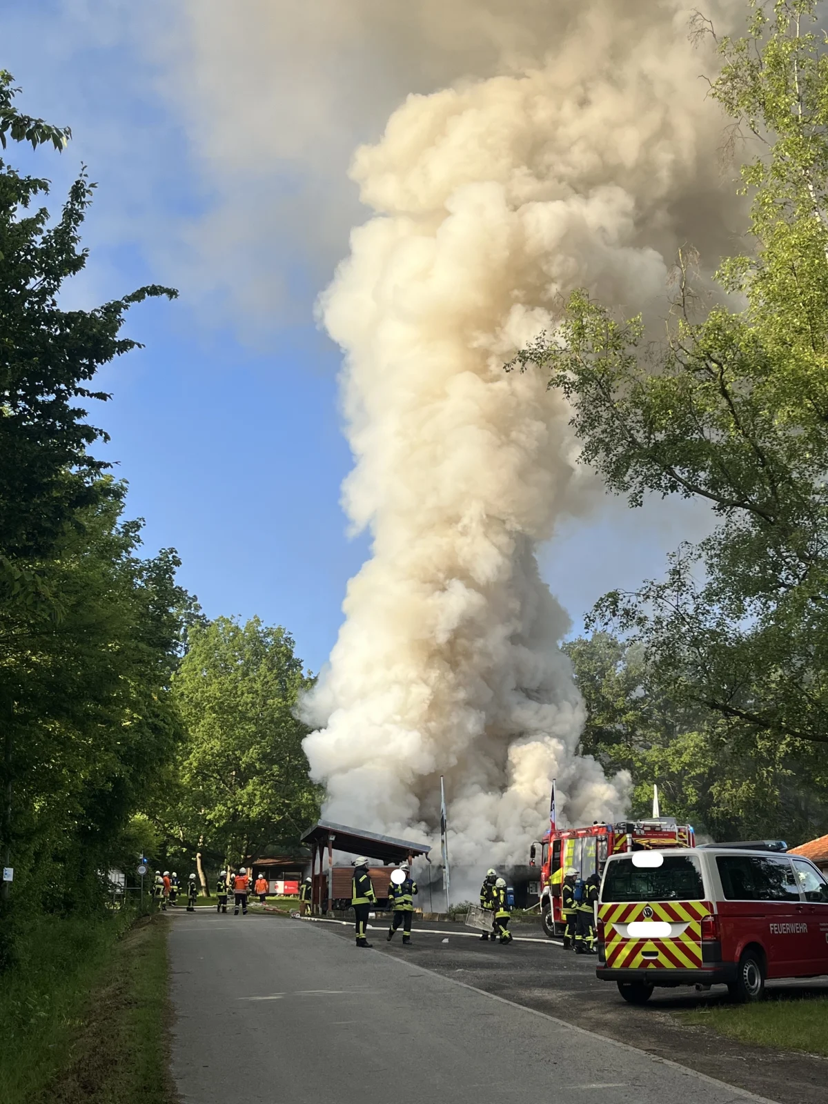 Dichter Rauch steigt von einer brennenden Lagerhalle auf, während Feuerwehrleute vor Ort den Brand bekämpfen. Fahrzeuge der Feuerwehr sind ebenfalls sichtbar.