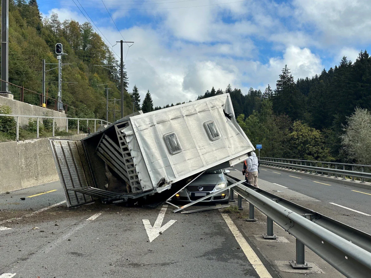 Ein umgestürzter Anhänger liegt auf einem grauen VW, blockiert die Straße neben einer Bahnlinie und einem Wald.