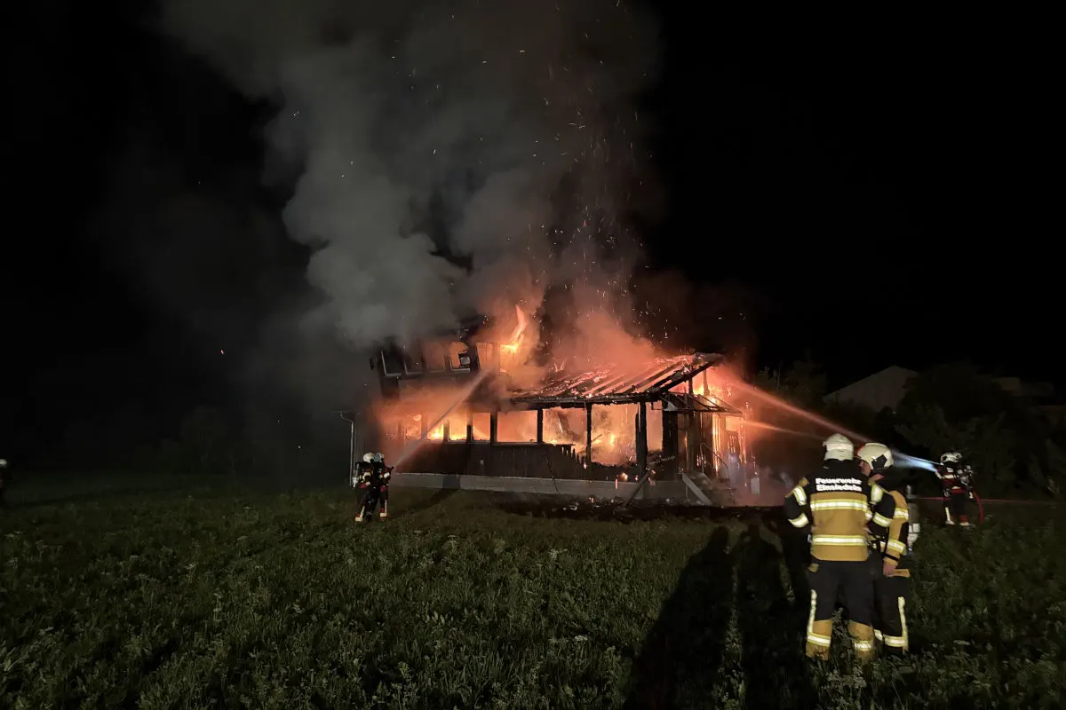 Mehrere Feuerwehrleute bekämpfen intensiv einen großen Brand in einem Holzgebäude mit Schläuchen. Rauch und Flammen steigen in die Nacht empor.
