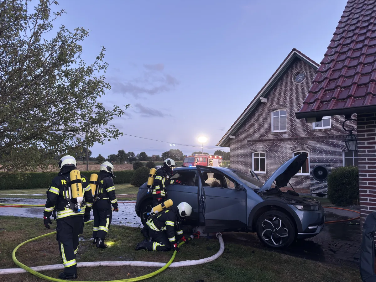 Mehrere Feuerwehrleute in Schutzkleidung arbeiten an einem Elektrofahrzeug mit geöffneter Motorhaube vor einem Ziegelhaus in ländlicher Umgebung.