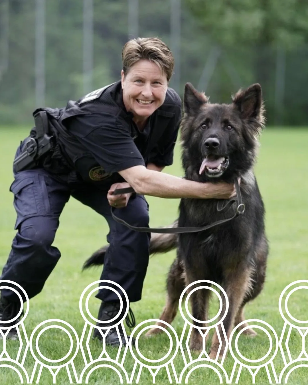 Ein lächelnder Polizeibeamter kniet auf einer grünen Wiese neben einem deutschen Schäferhund. Beide wirken aufmerksam.