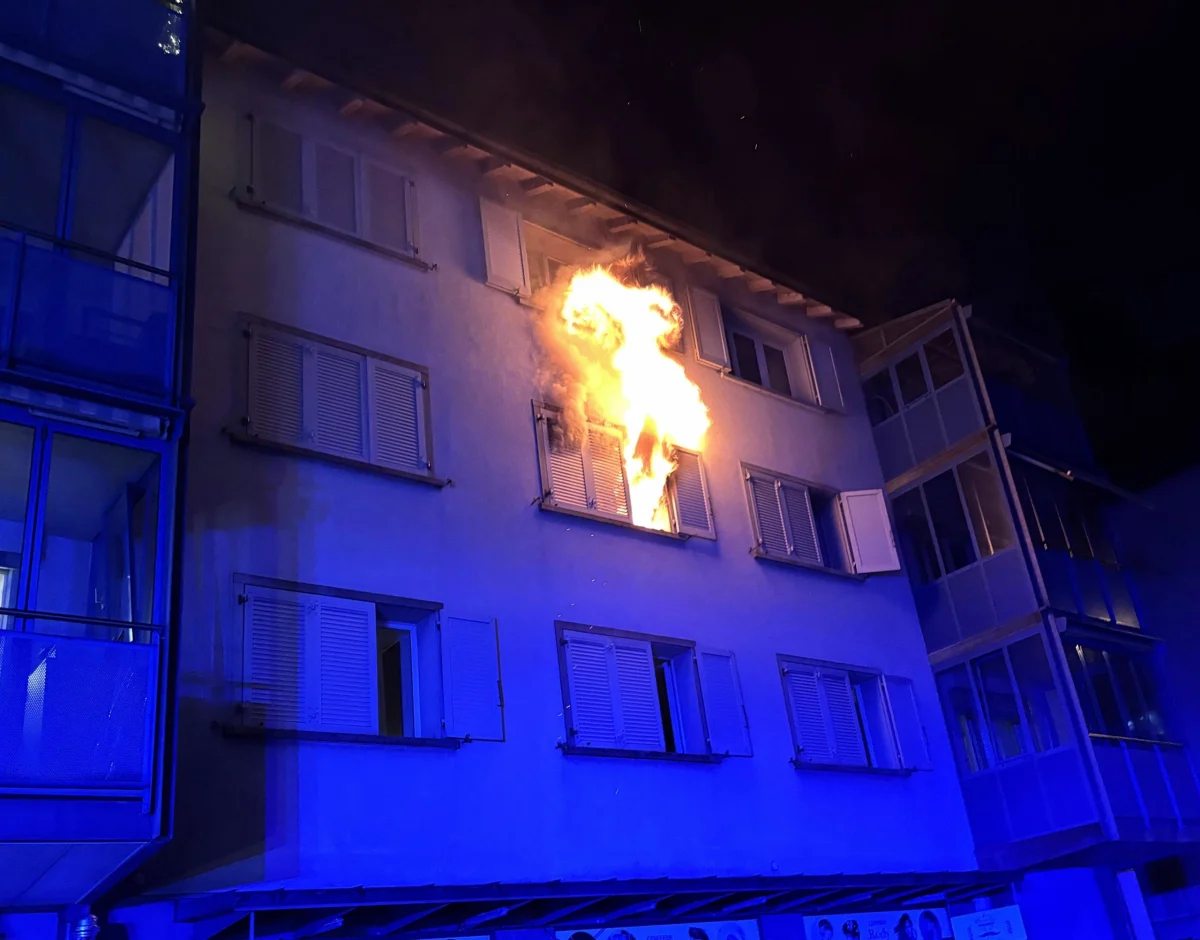 Ein oberes Stockwerk eines Wohngebäudes brennt, Flammen schlagen aus einem Fenster. Nachtaufnahme, blaues Licht erhellt die Fassade.