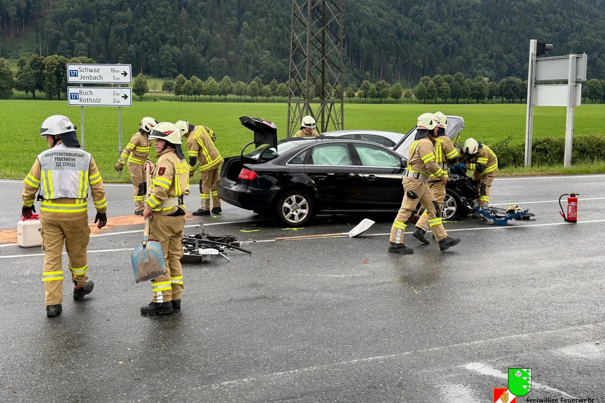 Mehrere Feuerwehrleute in Uniform arbeiten um ein beschädigtes, schwarzes Auto auf einer Landstraße. Im Hintergrund sind grüne Wiesen und Wald zu sehen.