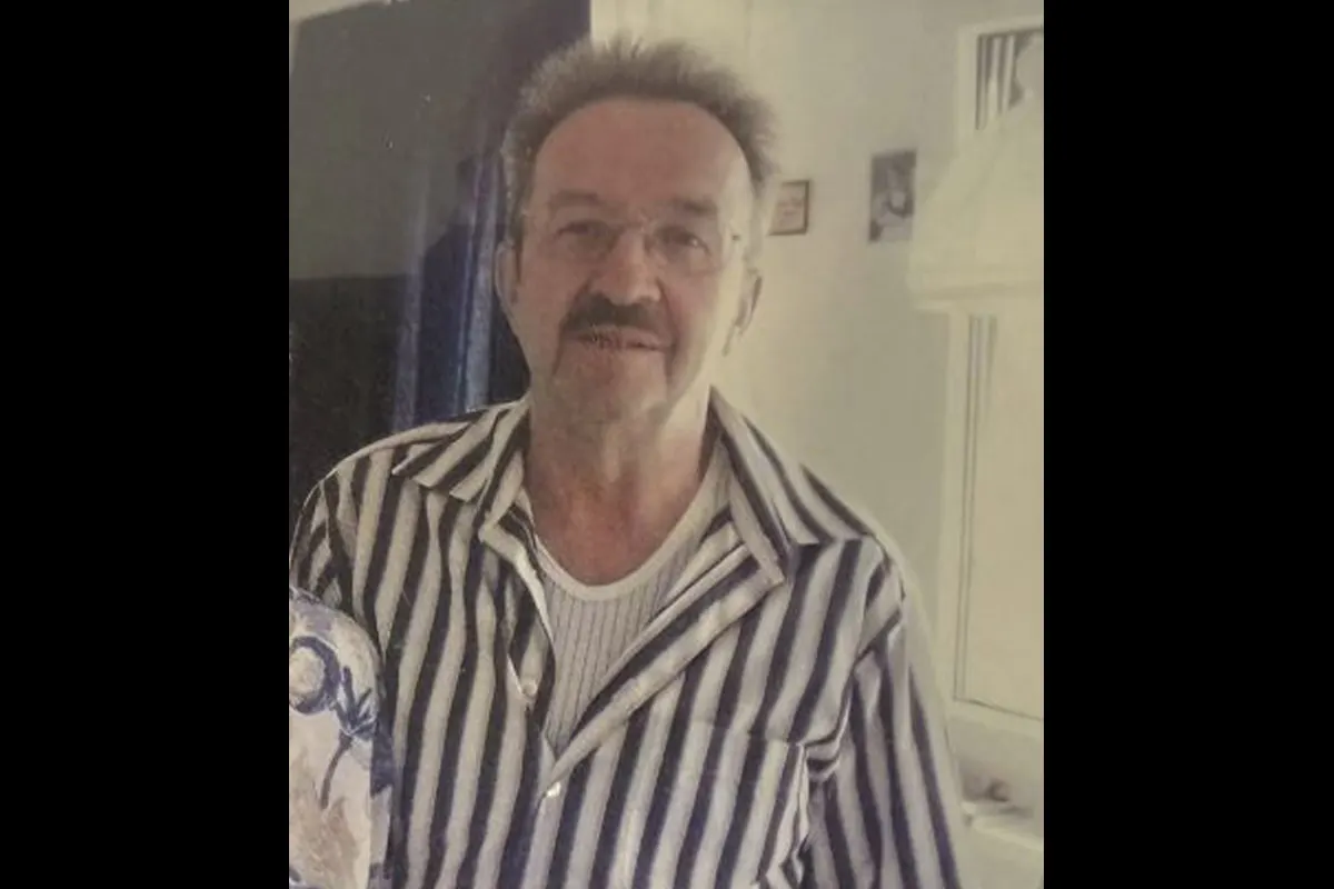 Ein älterer Mann mit gestreiftem Hemd und Bart blickt direkt in die Kamera, während er in einem Innenraum steht.