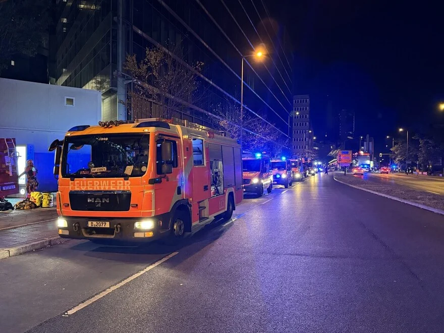 Ein Feuerwehrauto steht bei Nacht auf einer Straße, flankiert von weiteren Einsatzfahrzeugen. Bürogebäude sind im Hintergrund sichtbar.
