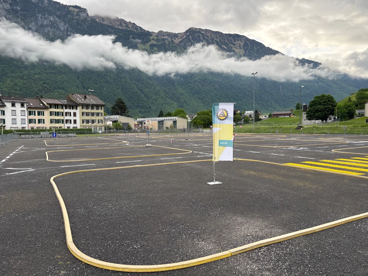 Ein Fahrsicherheitsplatz mit gelben Markierungen, eine Fahne mit Logo und Bergen im Hintergrund unter bewölktem Himmel.