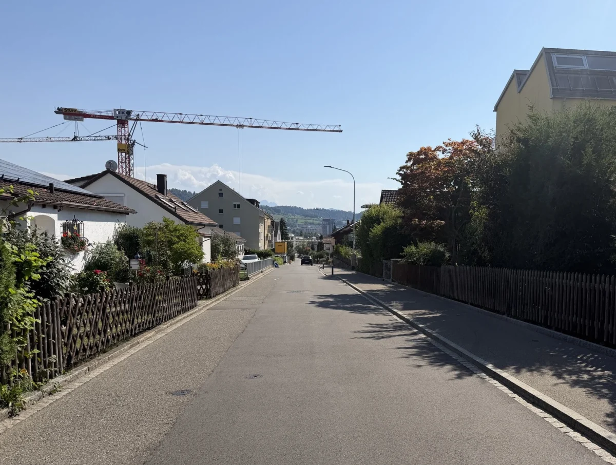 Eine leere, asphaltierte Straße in einem Wohngebiet. Links und rechts stehen Wohnhäuser, im Hintergrund ein Baukran.