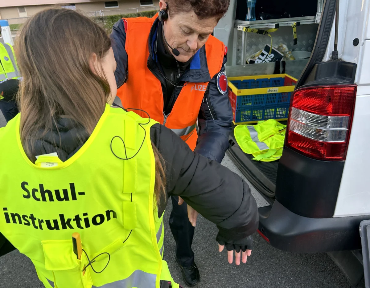 Eine Polizistin in orangefarbener Weste assistiert einem Kind, das eine gelbe Weste mit 'Schul- instruktion' trägt, bei einer Übung vor einem Fahrzeug.