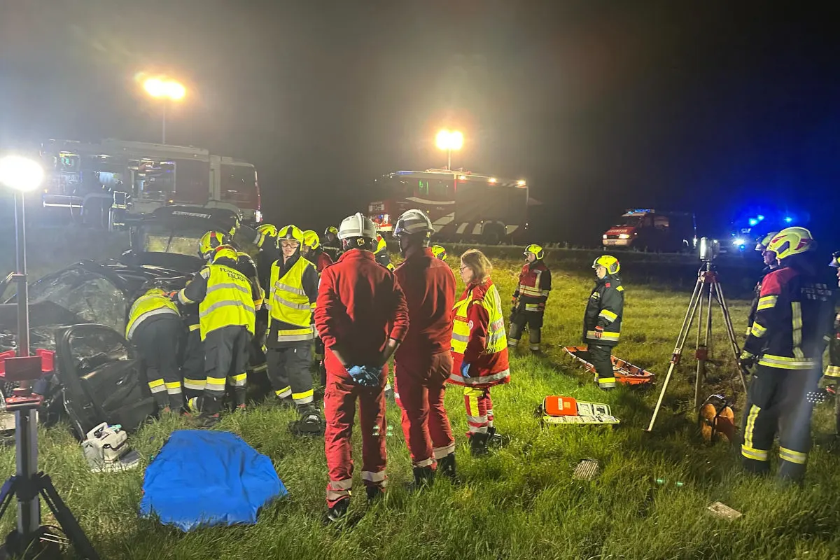 Mehrere Feuerwehrleute in reflektierenden Westen und Helmen arbeiten an einem beschädigten Fahrzeug bei Nacht. Im Hintergrund stehen beleuchtete Einsatzfahrzeuge.