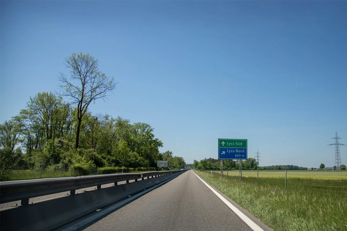 Eine leere Autobahn führt geradeaus, flankiert von Bäumen und einem Straßenschild, das nach Lyss-Süd und Lyss-Nord weist. Der Himmel ist klar.