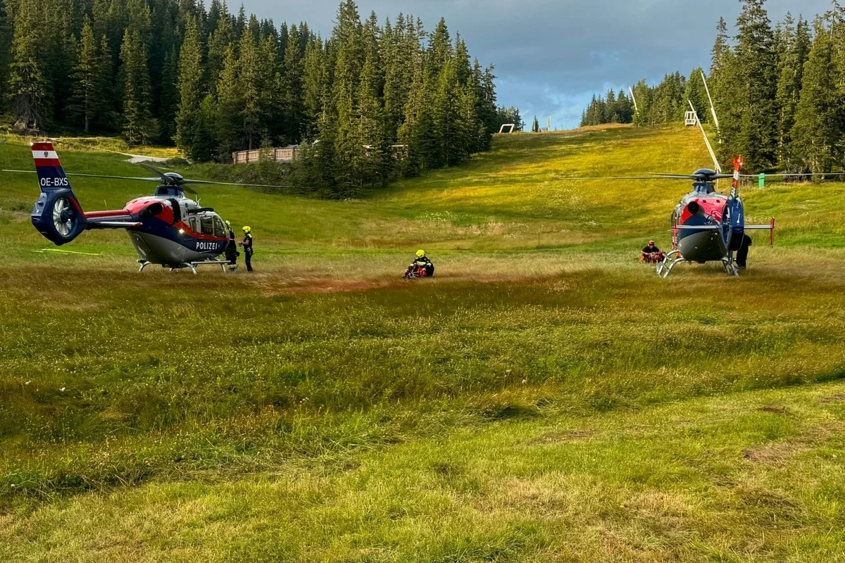 Zwei Polizeihubschrauber stehen auf einer grünen Wiese, umgeben von Bäumen in einer alpinen Landschaft. Mehrere Personen sind in der Nähe der Hubschrauber zu sehen.