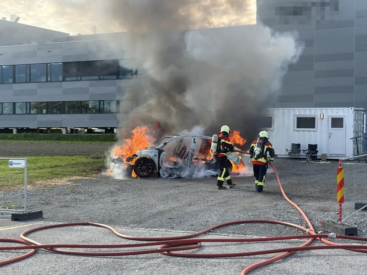 Ein Auto steht in Flammen vor einem modernen Bürogebäude. Zwei Feuerwehrleute in Schutzanzügen löschen den Brand.
