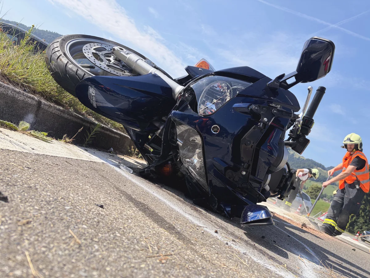 Ein umgestürztes Motorrad liegt auf der Straße, während Feuerwehrleute im Hintergrund arbeiten, um den Unfallort zu sichern.