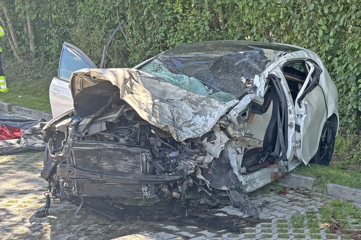 Ein stark beschädigtes weißes Auto steht auf einem gepflasterten Weg. Es hat erheblichen Frontschaden mit gebrochenen Teilen und zerbrochenen Scheiben.
