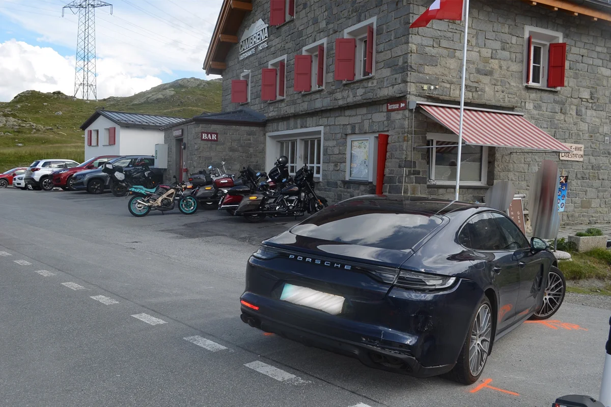 Ein schwarzer Porsche ist vor einem steinernen Gebäude mit roten Fensterläden und einer Schweizer Fahne geparkt. Mehrere Motorräder und Autos sind am Rand sichtbar.