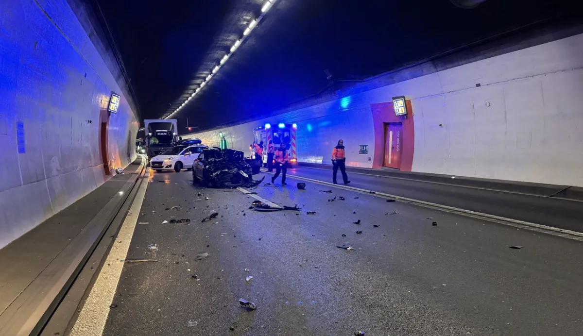 Ein stark beschädigtes Auto steht im Tunnel, umgeben von Trümmern und Einsatzkräften vor einem Notausgang.