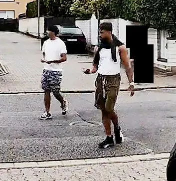 Zwei Männer gehen auf einer Straße in einer Wohngegend. Sie sind lässig gekleidet und scheinen in ein Gespräch vertieft zu sein.