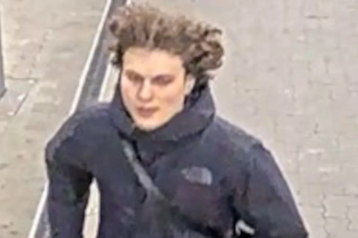 Ein Mann mit mittellangem, wehendem Haar trägt eine dunkle Jacke und eine Umhängetasche, während er auf einem Gehweg läuft.