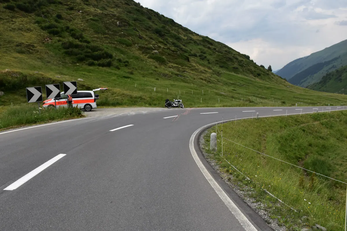 Ein weißes Polizeifahrzeug mit orangen Streifen steht am Straßenrand einer kurvigen Bergstraße. Dahinter ist ein Motorrad zu sehen.