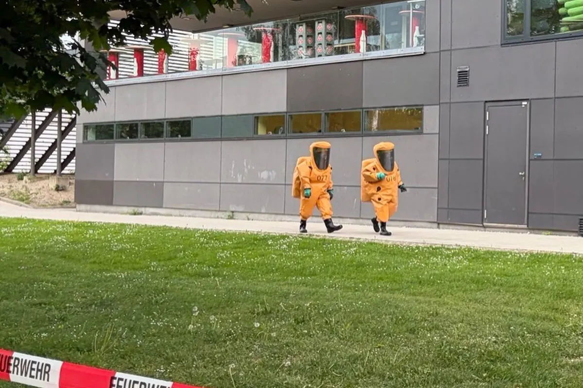 Zwei Personen in orangenen Schutzanzügen gehen auf einem Gehweg neben einem modernen Gebäude. Im Vordergrund ist eine Feuerwehrabsperrung zu sehen.