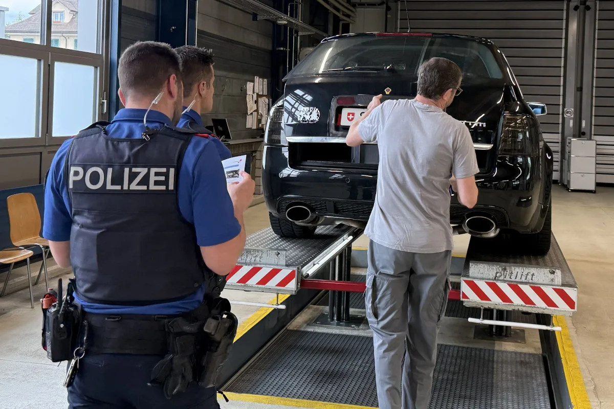 Zwei Polizisten stehen in einer Werkstatt vor einem schwarzen Auto, während ein Techniker das Auto überprüft. Das Auto ist auf einer Hebebühne angehoben.