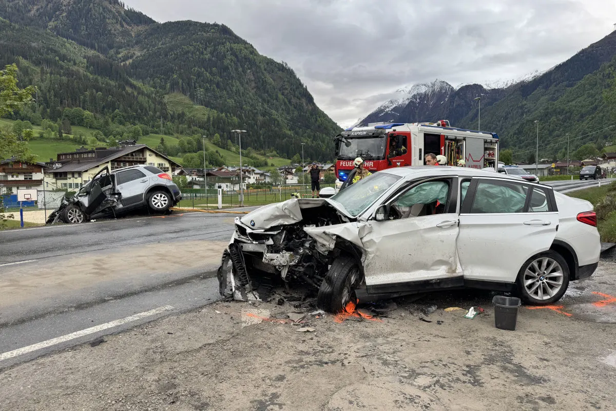 Zwei stark beschädigte Autos stehen nach einem Unfall auf einer Landstraße. Im Hintergrund sind Berge und ein Feuerwehrfahrzeug sichtbar.