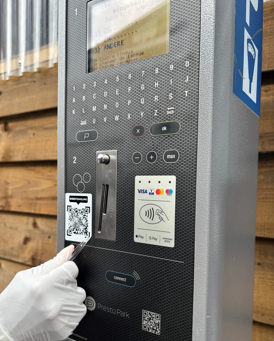 Ein Parkautomat wird von einer Person mit Handschuhen bedient, die einen QR-Code abtastet. Der Automat zeigt diverse Zahlungsmethoden.