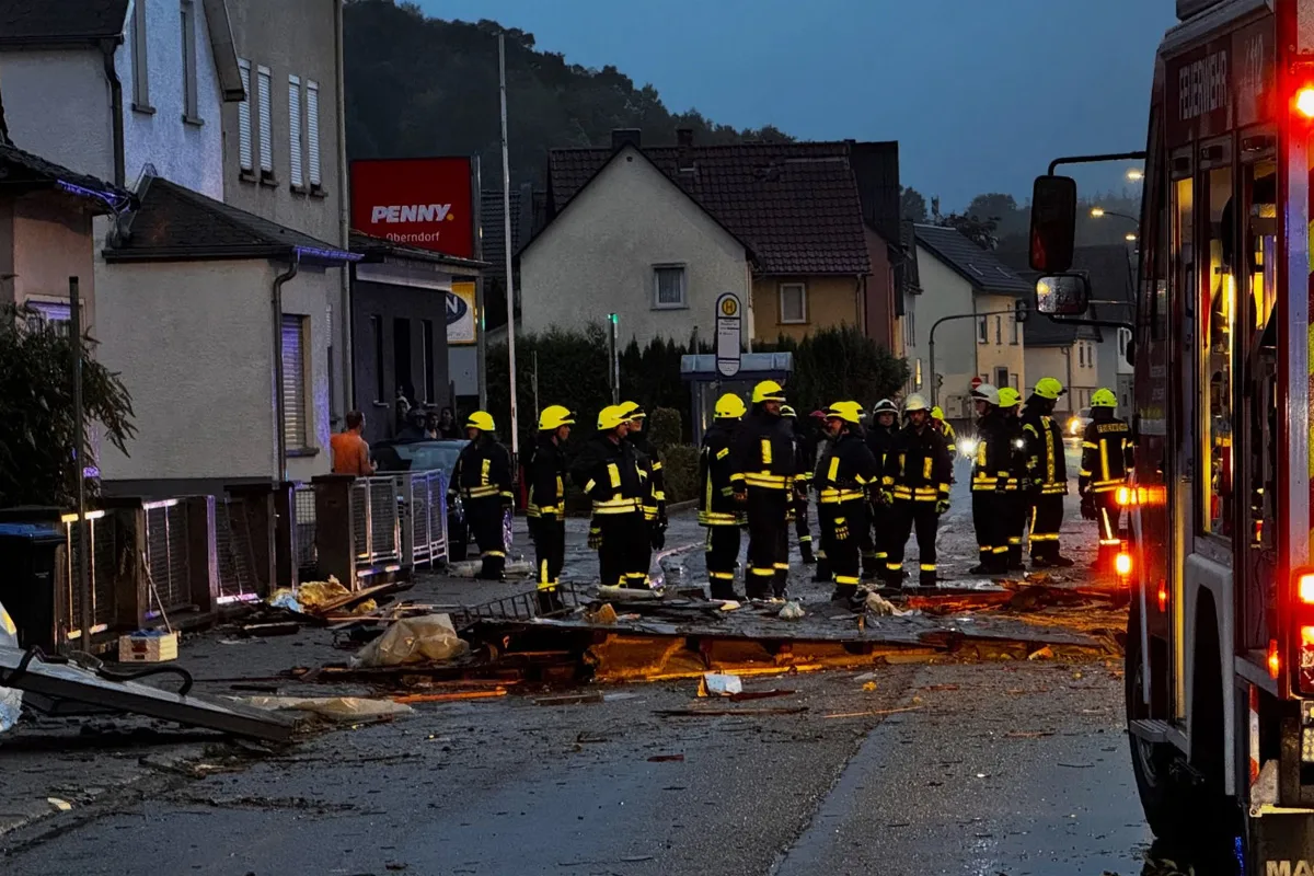 Mehrere Feuerwehrleute in Schutzkleidung stehen auf einer mit Trümmern übersäten Straße in einem Wohngebiet. Ein Feuerwehrfahrzeug mit eingeschaltetem Licht ist am rechten Bildrand sichtbar.