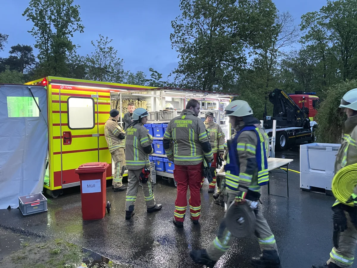 Feuerwehrleute stehen bei einem gelben Einsatzfahrzeug mit geöffneter Seitenklappe. Der Boden ist nass und es ist abends.