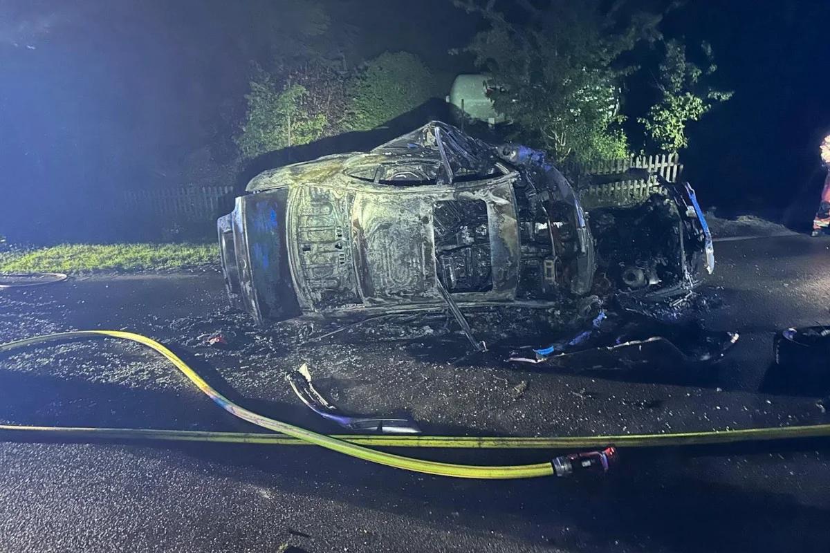 Ein stark beschädigtes und verbranntes Auto liegt auf der Seite auf einer Asphaltstraße. Die Szene ist nachts beleuchtet, umgeben von Bäumen und einem Zaun, mit einem Feuerwehrschlauch auf dem Boden.