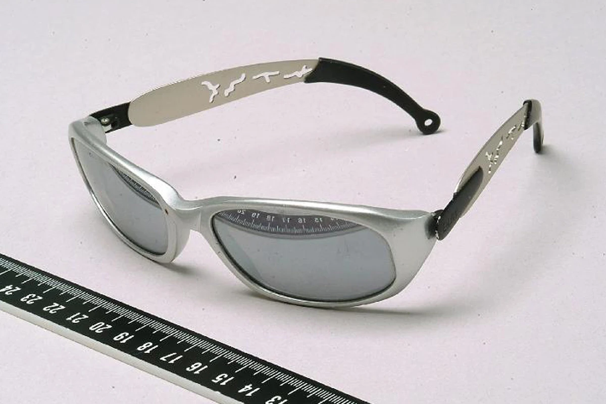 Eine silberne Sonnenbrille liegt auf einer Oberfläche, daneben ein Lineal zur Größenbestimmung. Die Brille hat dunkle Gläser und schwarze Bügel mit Mustern.