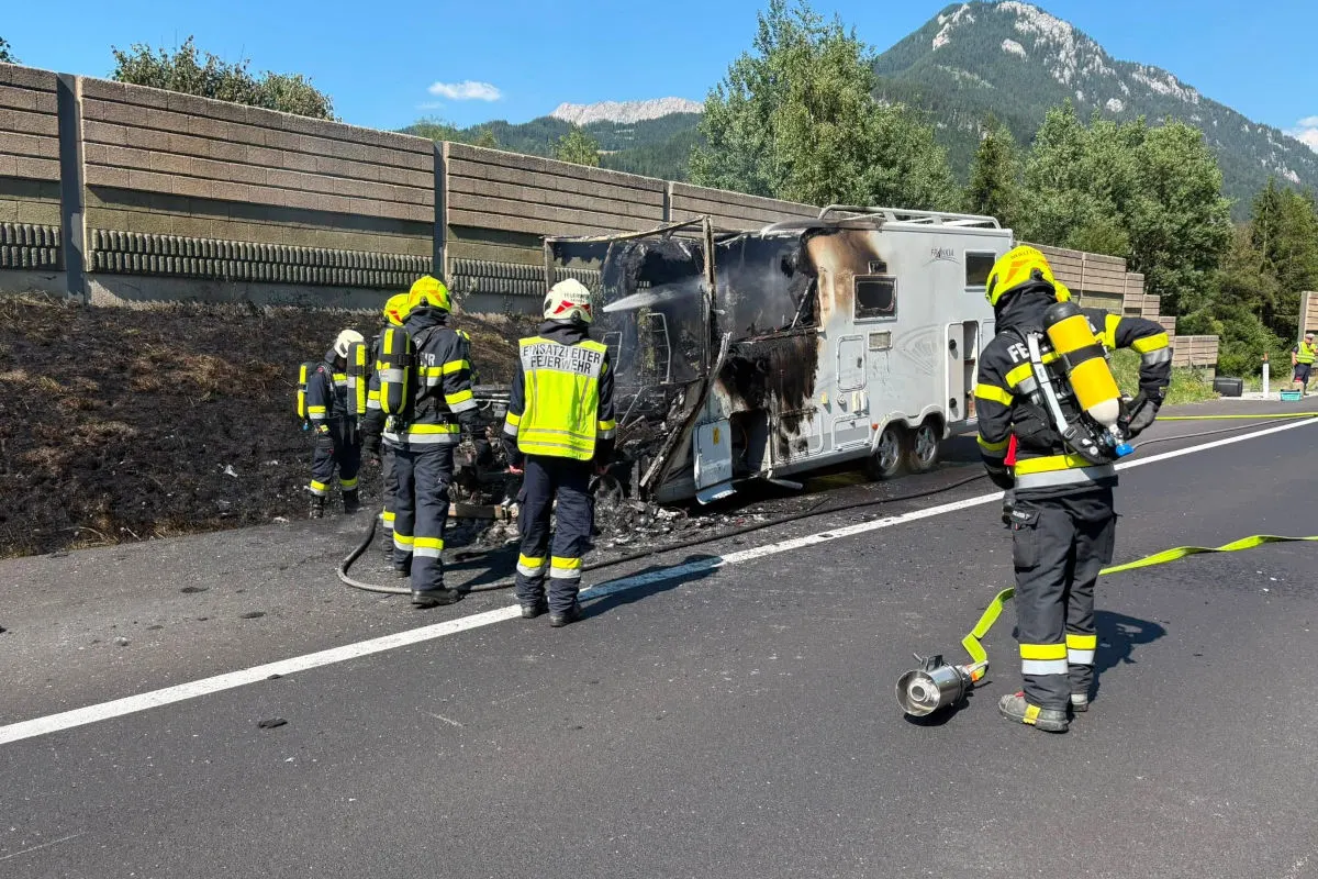 Mehrere Feuerwehrleute untersuchen die verkohlten Überreste eines Wohnwagens, der am Straßenrand steht. Der Hintergrund zeigt eine Lärmschutzwand und Berge unter klarem Himmel.