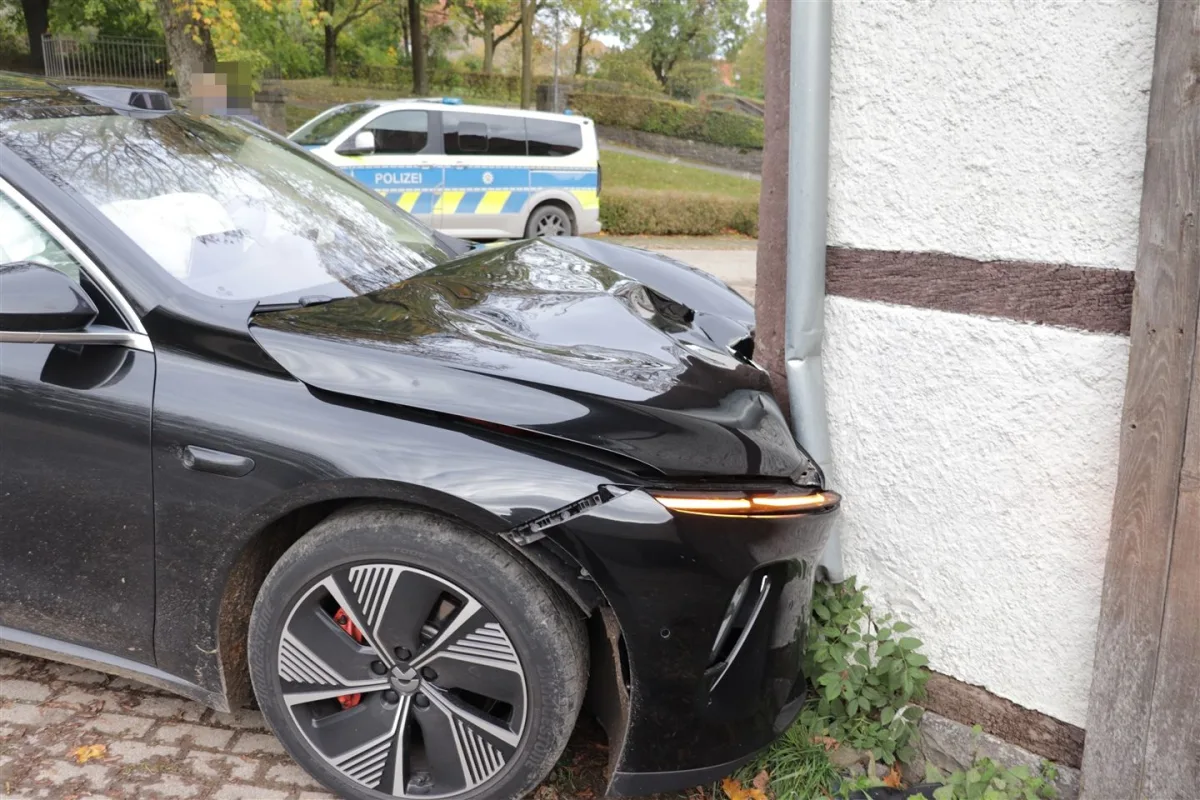 Ein schwarzes Auto ist mit der Vorderseite gegen eine weiße Mauer gefahren, ein Polizeifahrzeug steht im Hintergrund.