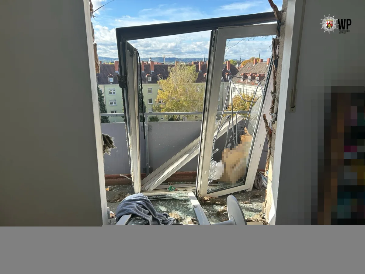 Das Bild zeigt ein stark beschädigtes Fenster in einem Gebäude, durch das eine Stadtlandschaft bei Tageslicht sichtbar ist. Die Fensterscheibe ist zersplittert, und die Rahmen sind stark verbogen.