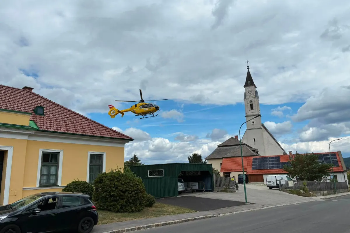 Ein gelber Rettungshubschrauber befindet sich in der Luft über einem Dorf. Im Vordergrund sind eine Kirche und mehrere Häuser sichtbar.