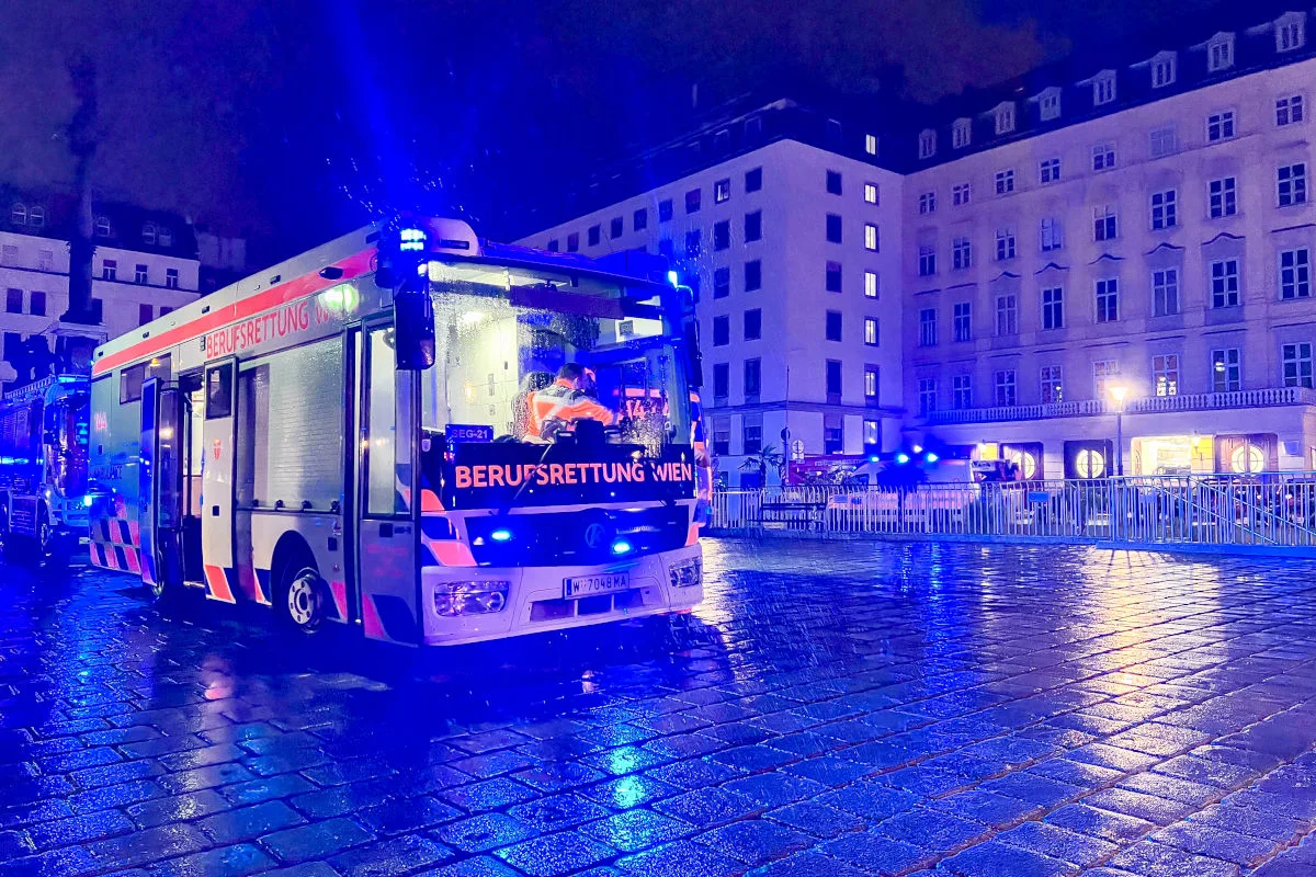 Ein Einsatzfahrzeug der Berufsrettung Wien steht bei Nacht auf einer nassen Straße in einem städtischen Bereich. Blaulicht beleuchtet die Szene, während umliegende Gebäude im Hintergrund sichtbar sind.