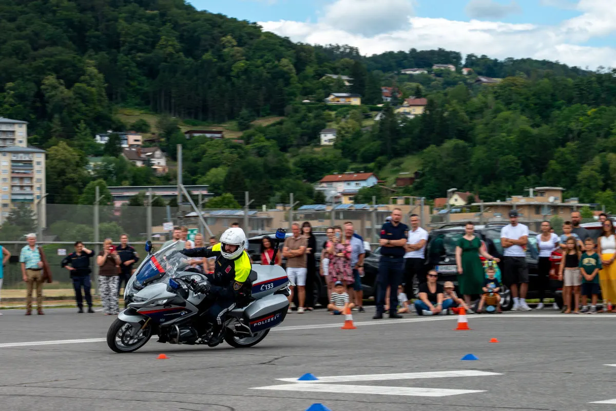 Ein Polizist auf einem Motorrad führt Manöver auf einer asphaltierten Fläche vor, während zahlreiche Menschen im Hintergrund zuschauen.