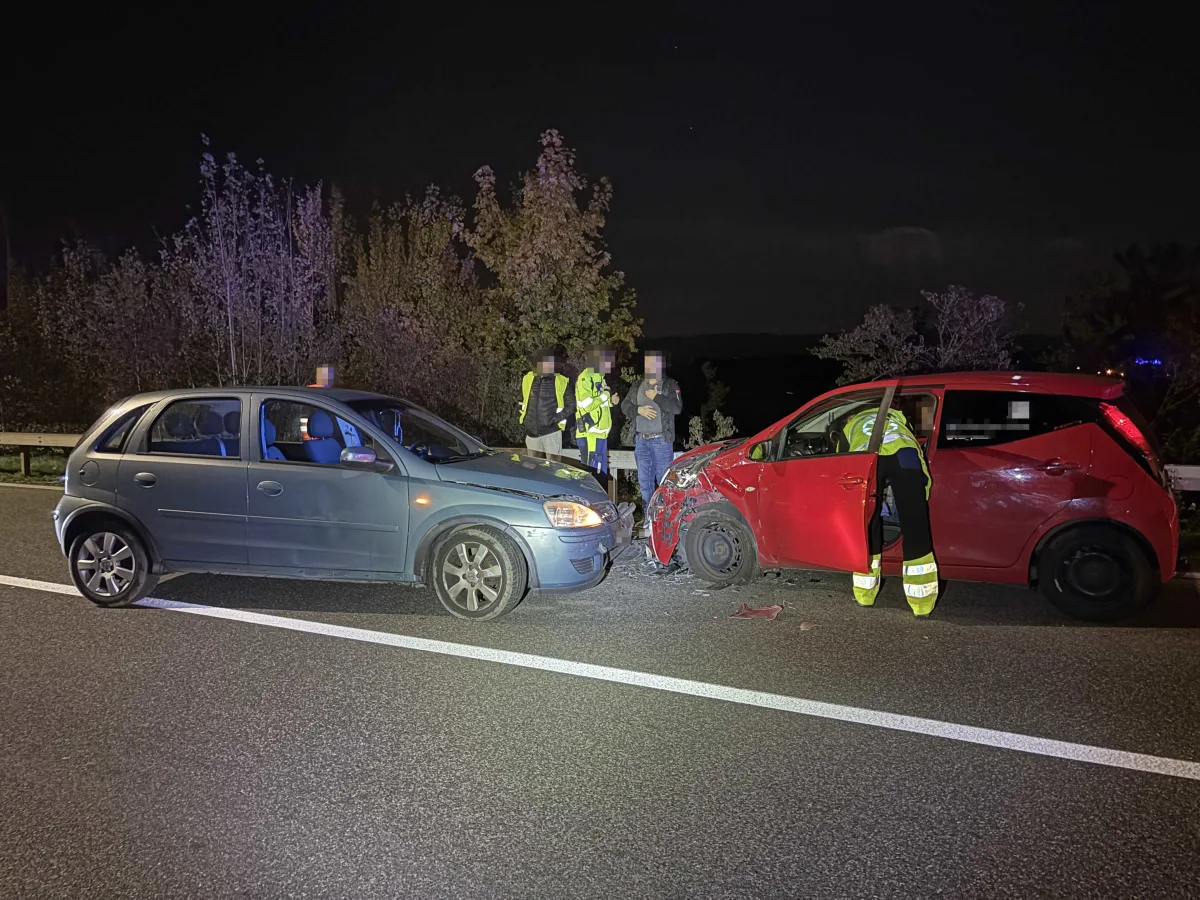 Zwei beschädigte Autos stehen frontal zueinander auf einer Straße, mehrere Personen stehen daneben. Es ist Nacht.