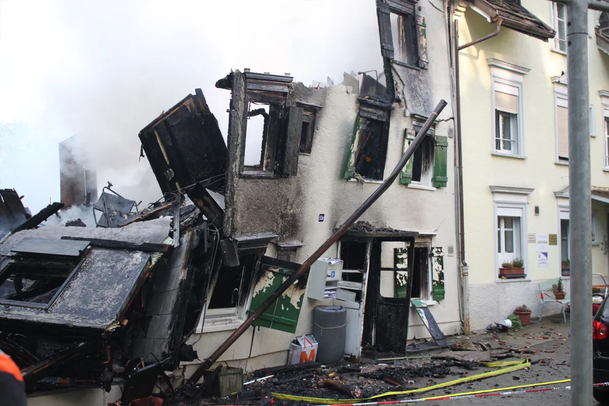 Ein Haus ist stark beschädigt, mit verbrannten Wänden und rauchender Zerstörung. Der vordere Teil des Gebäudes ist eingestürzt.