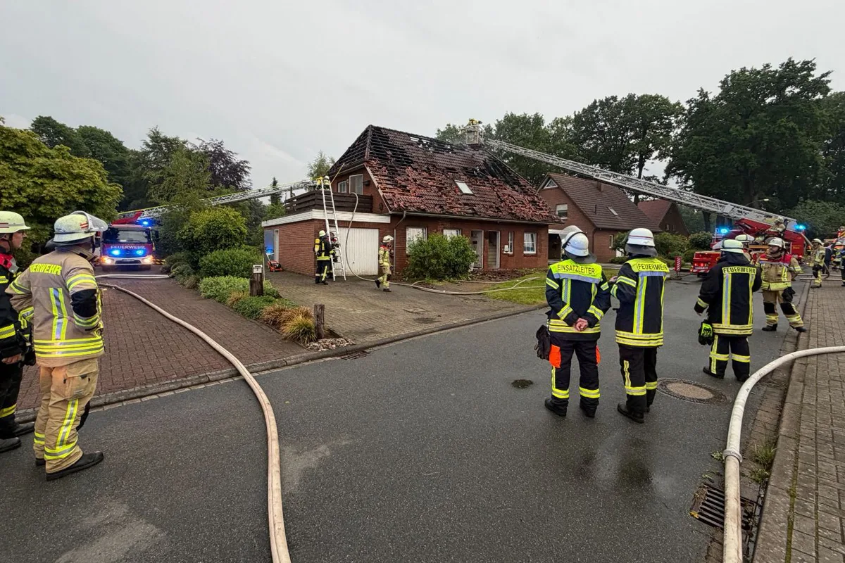 Mehrere Feuerwehrleute stehen vor einem Wohnhaus, dessen Dach durch einen Brand beschädigt wurde. Einsatzfahrzeuge im Hintergrund mit ausfahrbaren Leitern zur Brandbekämpfung.