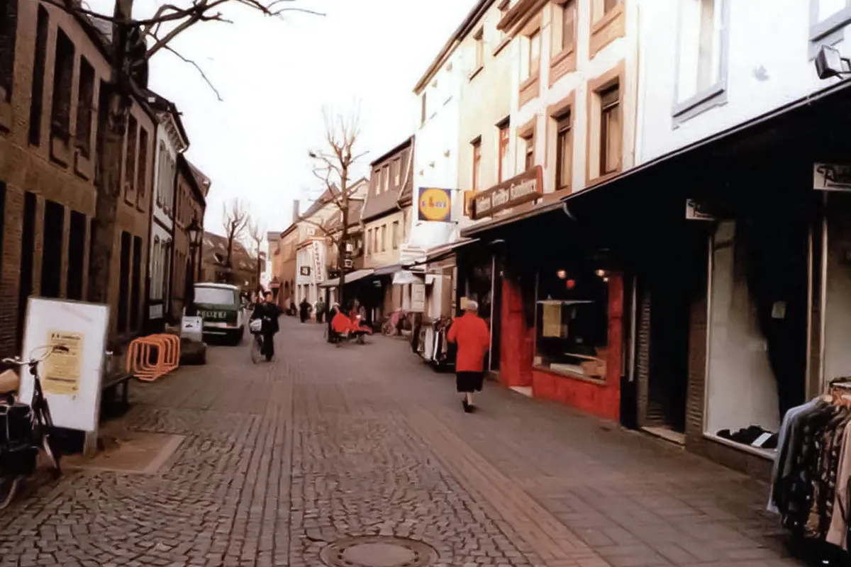 Eine belebte Fußgängerzone mit Geschäften, Passanten und einem geparkten Van auf Kopfsteinpflaster. Kleidung hängt an einem Ständer vor einem Geschäft.