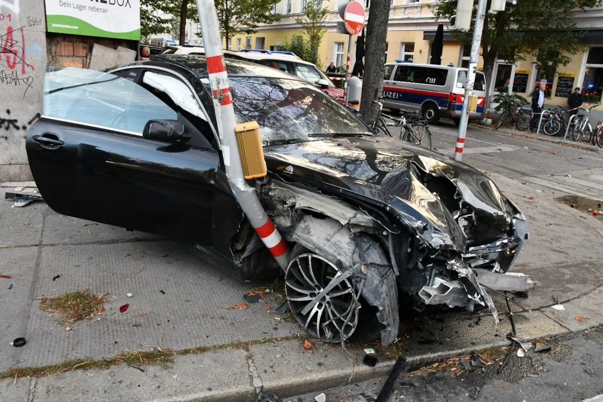 Ein schwarzes Auto mit erheblichen Frontschäden nach einer Kollision mit einem Lichtmast. Im Hintergrund sind Polizeifahrzeuge zu sehen.